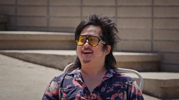 Movie still from “How It Ends” (2021), directed by Daryl Wein – A man with long black hair wearing sunglasses and sitting in a chair; Close Up shot, Low angle
