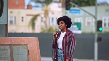 Movie still from “How It Ends” (2021), directed by Daryl Wein – A woman holding a microphone while standing in front of an easel; Medium shot, Over the shoulder angle
