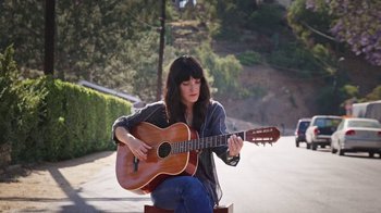 Movie still from “How It Ends” (2021), directed by Daryl Wein – A woman sitting on the side of a road playing a guitar; Medium shot, Low angle