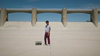 Movie still from “How It Ends” (2021), directed by Daryl Wein – A man standing next to a radio and a skateboard; Wide shot, Low angle