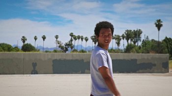 Movie still from “How It Ends” (2021), directed by Daryl Wein – A young man smiles while standing on a beach with palm trees in the background; Medium shot, Low angle