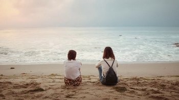 Movie still from “How It Ends” (2021), directed by Daryl Wein – Two women sitting on the beach looking out at the ocean; Wide shot, High angle