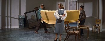 Movie still from “How to Marry a Millionaire” (1953), directed by Jean Negulesco – A man and a woman are standing next to a yellow couch; Wide shot, High angle