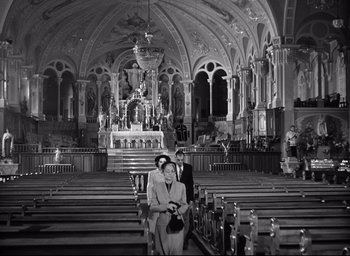 Movie still from “I Confess” (1953), directed by Alfred Hitchcock – Two people sitting in a large church with pedestals; Wide shot, High angle