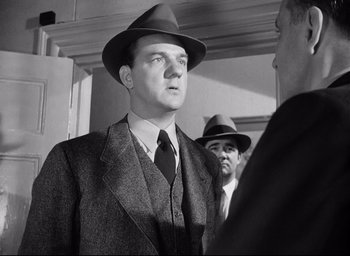 Movie still from “I Confess” (1953), directed by Alfred Hitchcock – A black and white photo of a man in a suit and hat; Medium shot, Low angle