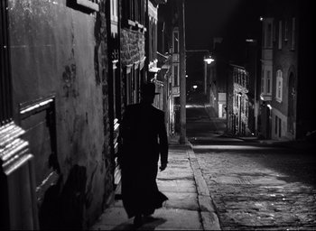 Movie still from “I Confess” (1953), directed by Alfred Hitchcock – A man walking down a street at night; Wide shot, Low angle