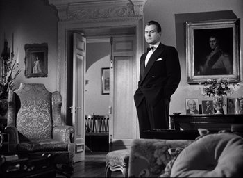 Movie still from “I Confess” (1953), directed by Alfred Hitchcock – A black and white photo of a man in a suit and bow tie; Medium shot, Low angle