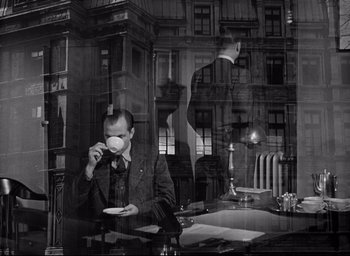 Movie still from “I Confess” (1953), directed by Alfred Hitchcock – A black and white photo of a man drinking a cup of coffee; Wide shot, Over the shoulder angle