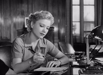 Movie still from “I Confess” (1953), directed by Alfred Hitchcock – A woman sitting at a desk writing on a piece of paper; Medium shot, Low angle