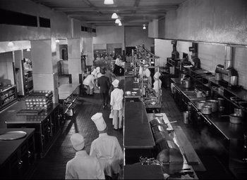 Movie still from “I Confess” (1953), directed by Alfred Hitchcock – A group of people standing in a kitchen preparing food; Wide shot, High angle