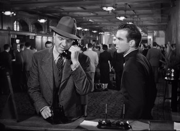 Movie still from “I Confess” (1953), directed by Alfred Hitchcock – A man talking on a phone while standing next to another man; Medium shot, Low angle
