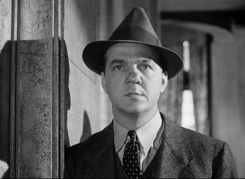 Movie still from “I Confess” (1953), directed by Alfred Hitchcock – A man wearing a suit and a hat is standing in front of a wall; Close Up shot, Low angle