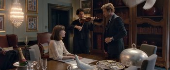 Movie still from “I Give It a Year” (2013), directed by Dan Mazer – A man playing a violin for a woman and another man standing at a table; Medium shot, Over the shoulder angle