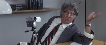 Movie still from “I Heart Huckabees” (2004), directed by David O. Russell – An older man sitting at a table with a camera; Medium shot, Over the shoulder angle