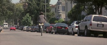 Movie still from “I Heart Huckabees” (2004), directed by David O. Russell – A woman riding a bike down a street next to parked cars; Wide shot, High angle