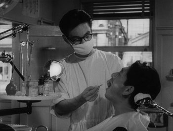 Movie still from “I Live in Fear” (1955), directed by Akira Kurosawa – A man in glasses and a mask is in a dentist's office; Medium shot, Over the shoulder angle