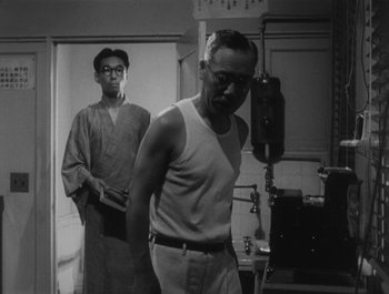 Movie still from “I Live in Fear” (1955), directed by Akira Kurosawa – A man standing next to another man in front of a microphone; Medium shot, Low angle
