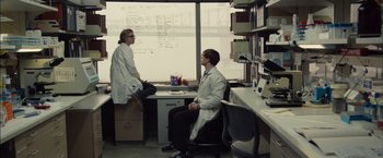 Movie still from “I Origins” (2014), directed by Mike Cahill – Two people in lab coats sitting at a desk in front of a window; Wide shot, Over the shoulder angle