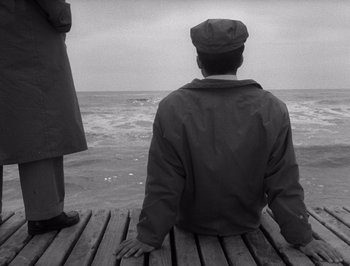 Movie still from “I Vitelloni” (1953), directed by Federico Fellini – A man sitting on top of a wooden dock; Medium shot, High angle
