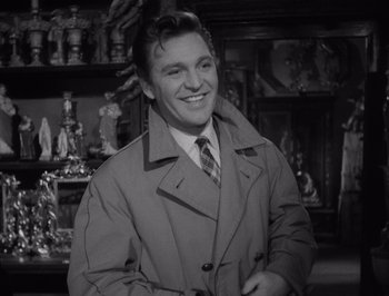 Movie still from “I Vitelloni” (1953), directed by Federico Fellini – A man in a trench coat and a tie smiling for the camera; Medium shot, Over the shoulder angle