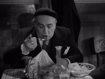 Movie still from “I Vitelloni” (1953), directed by Federico Fellini – An older man sitting at a table eating a piece of food; Medium shot, High angle