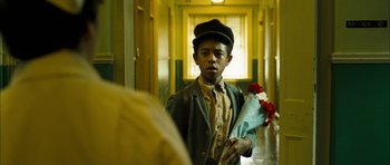 Movie still from “I'm Not There” (2007), directed by Todd Haynes – A young boy holding a bouquet of flowers in a hallway; Close Up shot, Over the shoulder angle