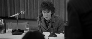 Movie still from “I'm Not There” (2007), directed by Todd Haynes – A person sitting at a table with a microphone; Close Up shot, Over the shoulder angle