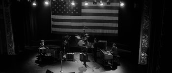Movie still from “I'm Not There” (2007), directed by Todd Haynes – A group of people standing in front of an american flag; Extreme Wide shot, High angle
