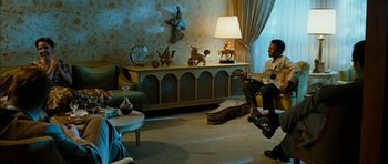 Movie still from “I'm Not There” (2007), directed by Todd Haynes – A man sitting in a living room holding a guitar; Wide shot, Over the shoulder angle