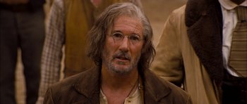 Movie still from “I'm Not There” (2007), directed by Todd Haynes – An older man with long grey hair wearing glasses and a brown jacket; Close Up shot, Over the shoulder angle