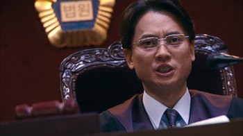 Movie still from “I'm a Cyborg, But That's OK” (2006), directed by Park Chan-wook – A man wearing glasses and a suit is sitting in front of a chair; Close Up shot, Low angle