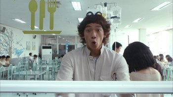 Movie still from “I'm a Cyborg, But That's OK” (2006), directed by Park Chan-wook – A young man is sitting at a table in front of a bunch of chairs; Medium shot, Low angle