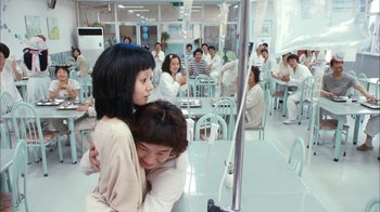 Movie still from “I'm a Cyborg, But That's OK” (2006), directed by Park Chan-wook – Two women hugging in a hospital room with a group of people in the background; Wide shot, Over the shoulder angle