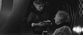 Movie still from “Voyage to the End of the Universe” (1963), directed by Jindrich Polák – An older woman serving a man at a party; Medium shot, Over the shoulder angle