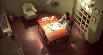 Movie still from “Il Mare” (2000), directed by Hyun-seung Lee – A woman sitting at a wooden table in a dimly - lit room; Extreme Wide shot, High angle