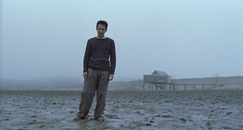 Movie still from “Il Mare” (2000), directed by Hyun-seung Lee – A man standing in the sand in front of a house; Wide shot, Low angle