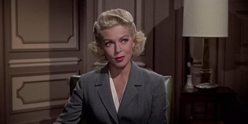 Movie still from “Imitation of Life” (1959), directed by Douglas Sirk – A woman with blonde hair sitting in a chair wearing a suit; Close Up shot, Low angle
