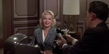 Movie still from “Imitation of Life” (1959), directed by Douglas Sirk – A woman in a gray suit is being interviewed by a man; Medium shot, Over the shoulder angle
