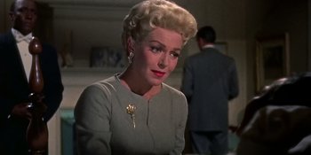 Movie still from “Imitation of Life” (1959), directed by Douglas Sirk – An older woman wearing a gray dress and a gold flower pin; Close Up shot, Over the shoulder angle
