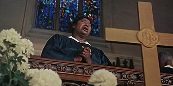 Movie still from “Imitation of Life” (1959), directed by Douglas Sirk – A man in a graduation cap is praying in front of a stained glass window; Medium shot, Low angle