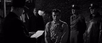 Movie still from “In Cold Blood” (1967), directed by Richard Brooks – A man chained to a brick wall while two other men watch; Medium shot, Over the shoulder angle