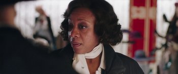 Movie still from “In Fabric” (2018), directed by Peter Strickland – An older woman wearing a black jacket and white neck scarf; Close Up shot, Over the shoulder angle