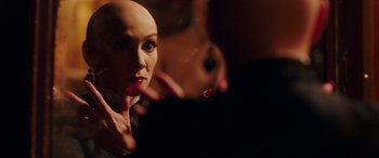 Movie still from “In Fabric” (2018), directed by Peter Strickland – A woman with a bald head is putting lipstick on her lips; Close Up shot, Over the shoulder angle