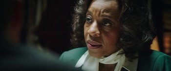 Movie still from “In Fabric” (2018), directed by Peter Strickland – An older woman with curly black hair wearing a green jacket; Close Up shot, Over the shoulder angle