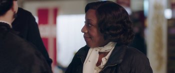 Movie still from “In Fabric” (2018), directed by Peter Strickland – An older woman wearing a black jacket and a white neck scarf; Close Up shot, Over the shoulder angle