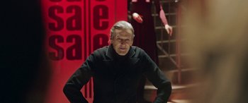 Movie still from “In Fabric” (2018), directed by Peter Strickland – An older man wearing a black jacket and standing in front of a red wall; Medium shot, Over the shoulder angle
