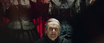 Movie still from “In Fabric” (2018), directed by Peter Strickland – An old man in a black shirt and a red and black dress; Close Up shot, Low angle