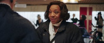 Movie still from “In Fabric” (2018), directed by Peter Strickland – A woman in a black jacket is smiling; Close Up shot, Over the shoulder angle