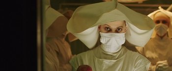 Movie still from “In Fabric” (2018), directed by Peter Strickland – A woman wearing a surgical mask while standing next to an apple; Medium shot, Low angle