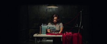 Movie still from “In Fabric” (2018), directed by Peter Strickland – A person sitting at a table with a sewing machine in front of him; Medium shot, High angle